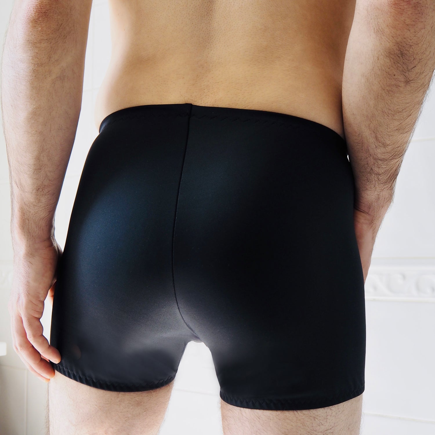 Back of a man's lower body wearing black swimwear trunks in a shower