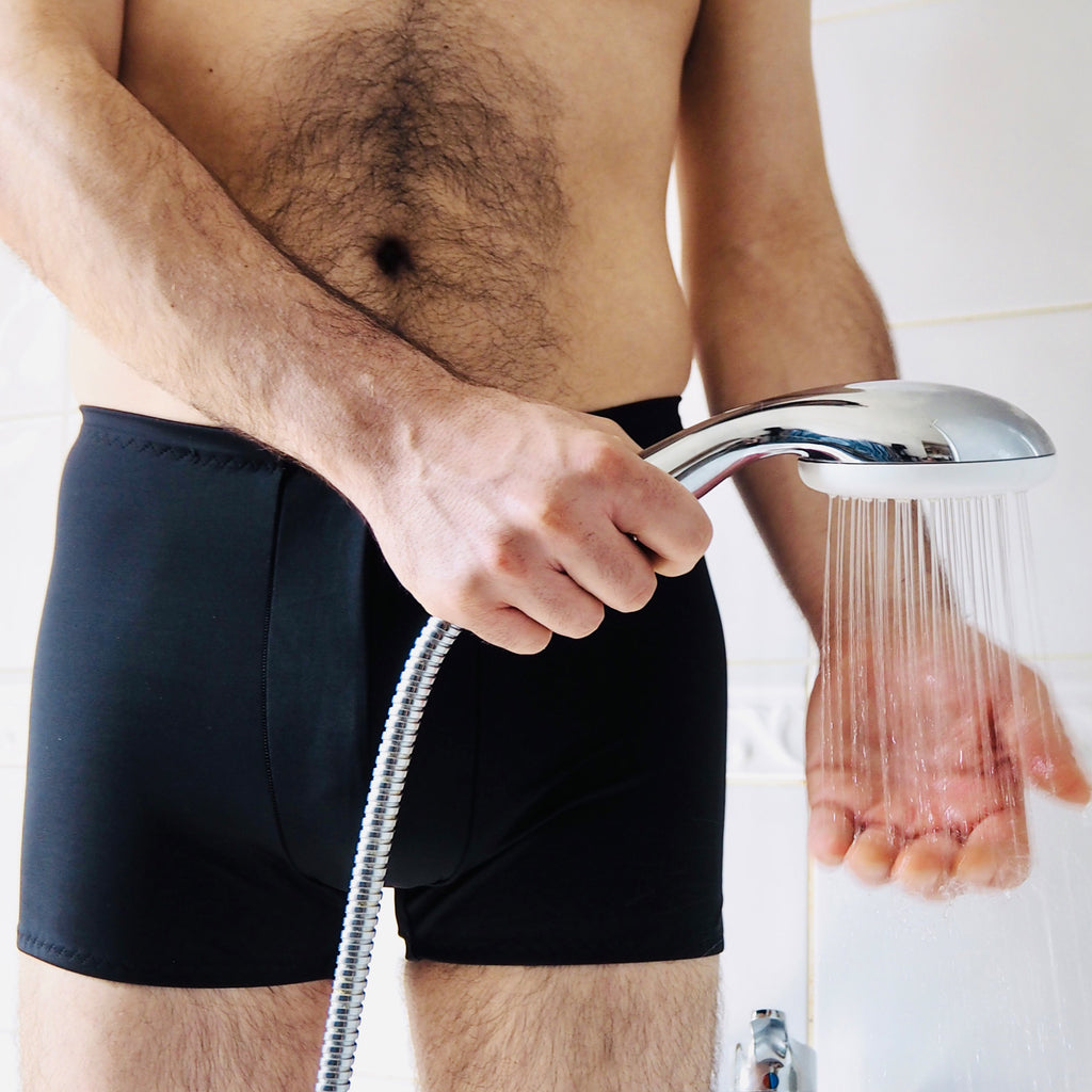 Lower body of a man with chest hair standing in a shower with black trunks holding a shower head ready to get wet