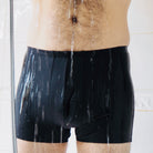 Lower body of a man who stands in the shower while water drips onto black trunks 